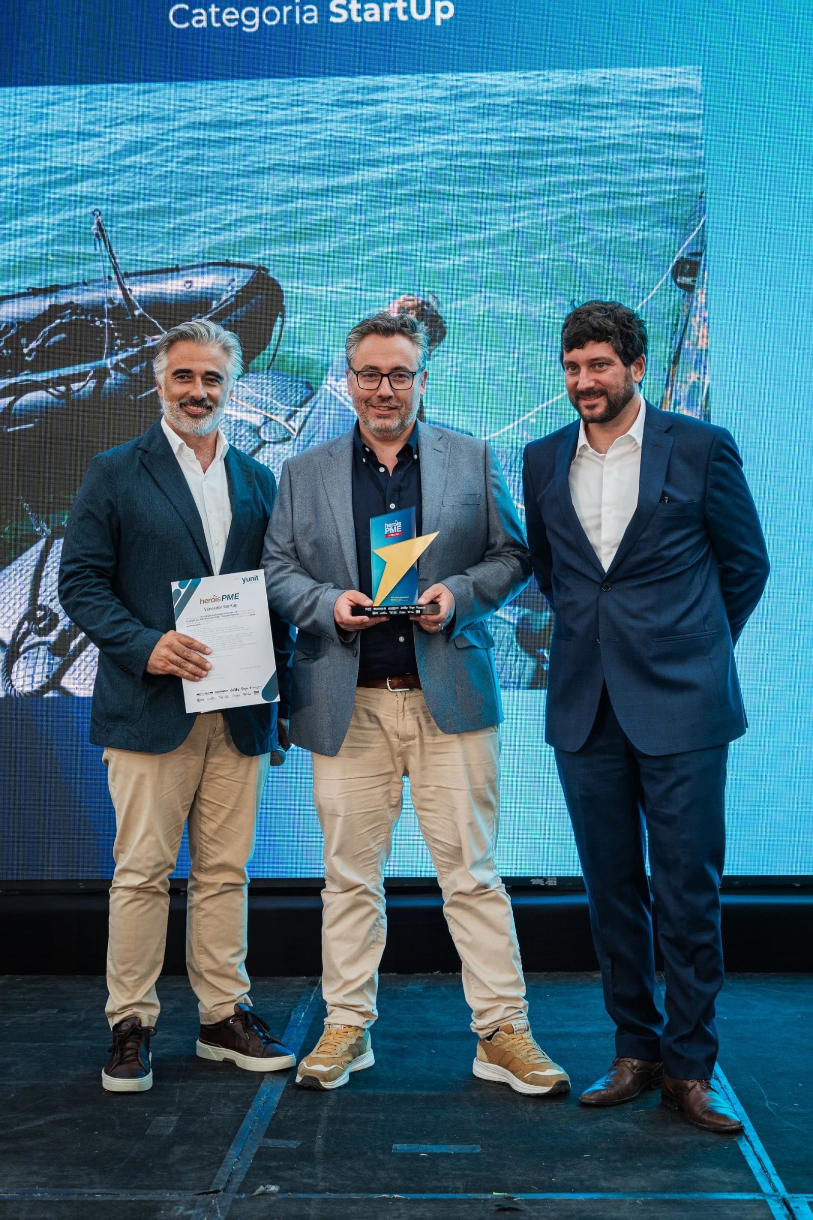 Três homens de fato num palco, dois segurando um troféu e um diploma, com ecrã atrás onde se lê "Categoria StartUp"