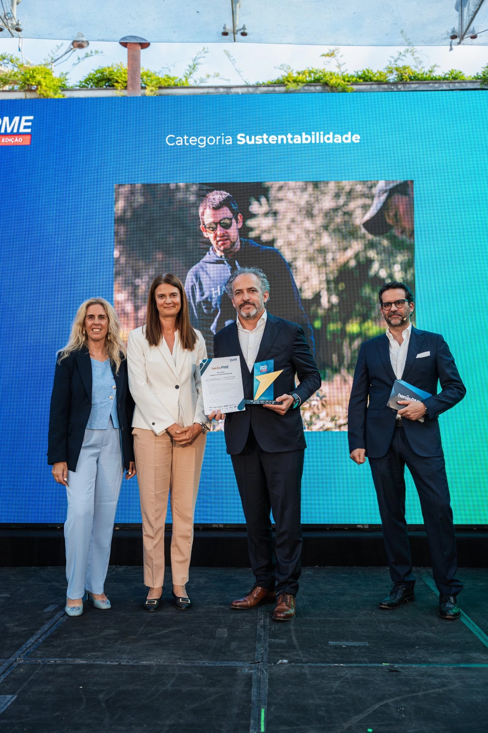 Quatro pessoas em palco; um homem segura diploma e troféu diante de ecrã com "Categoria Sustentabilidade"