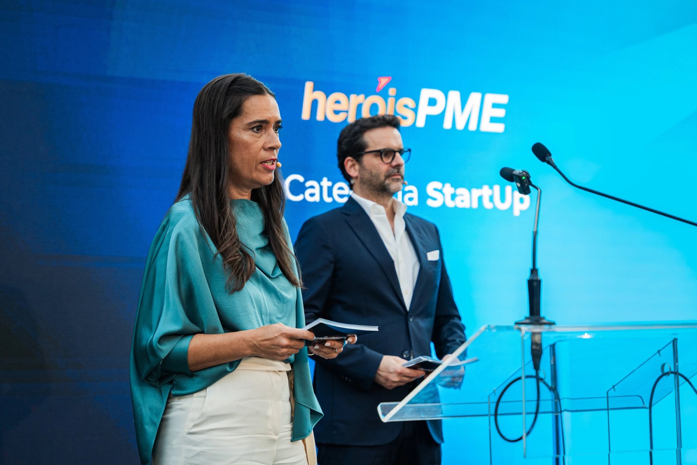 Mulher e homem em palco junto a um púlpito, com ecrã azul onde se lê "heróisPME" e "Categoria StartUp"